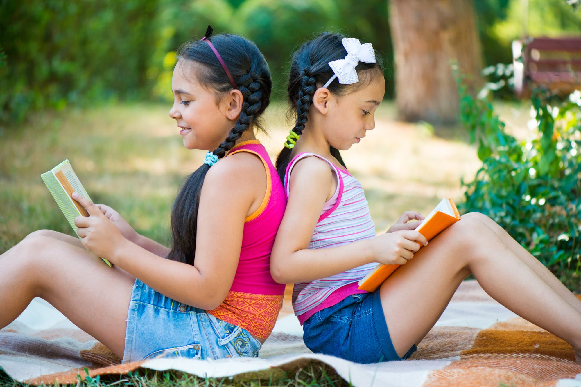 Two girls reading books outdoors on a blanket.